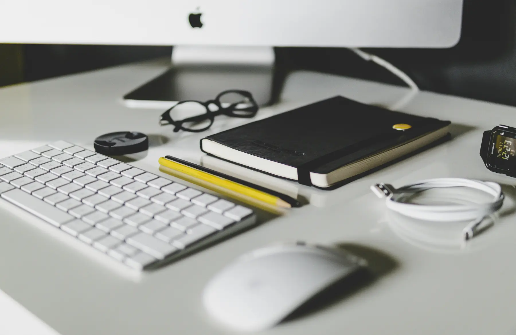 A clean white desk with an iMac, a keyboard and mouse, glasses, a notebook and a smartwatch.