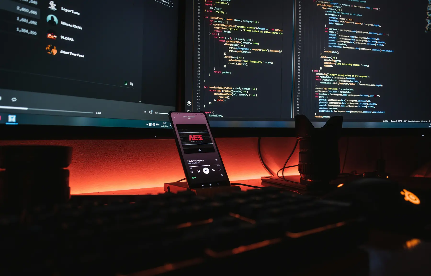 A dark desk setup at night: dual monitors with code and stats, a phone playing music, lit by an orange glow.