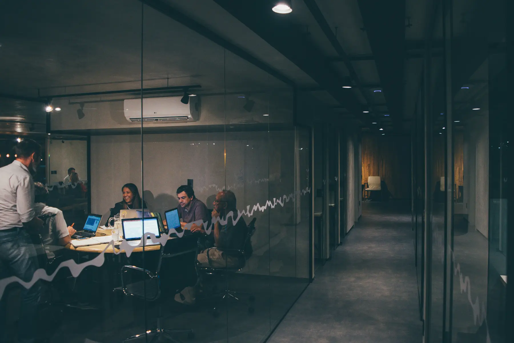 A team meeting around laptops in a glass-walled office at night, seen from the hallway.